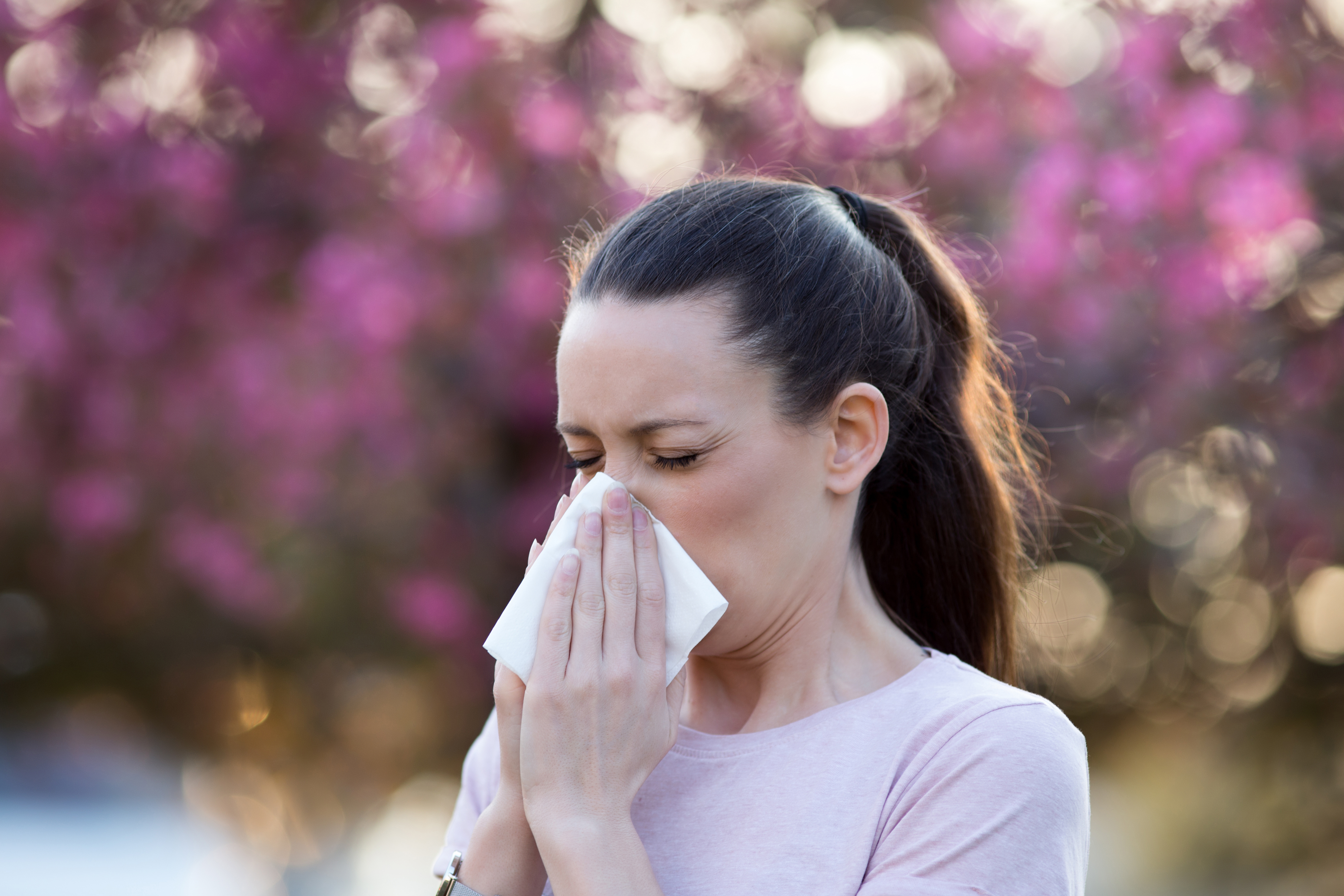 Young pretty woman blowing nose in front of blooming tree. Spring allergy concept